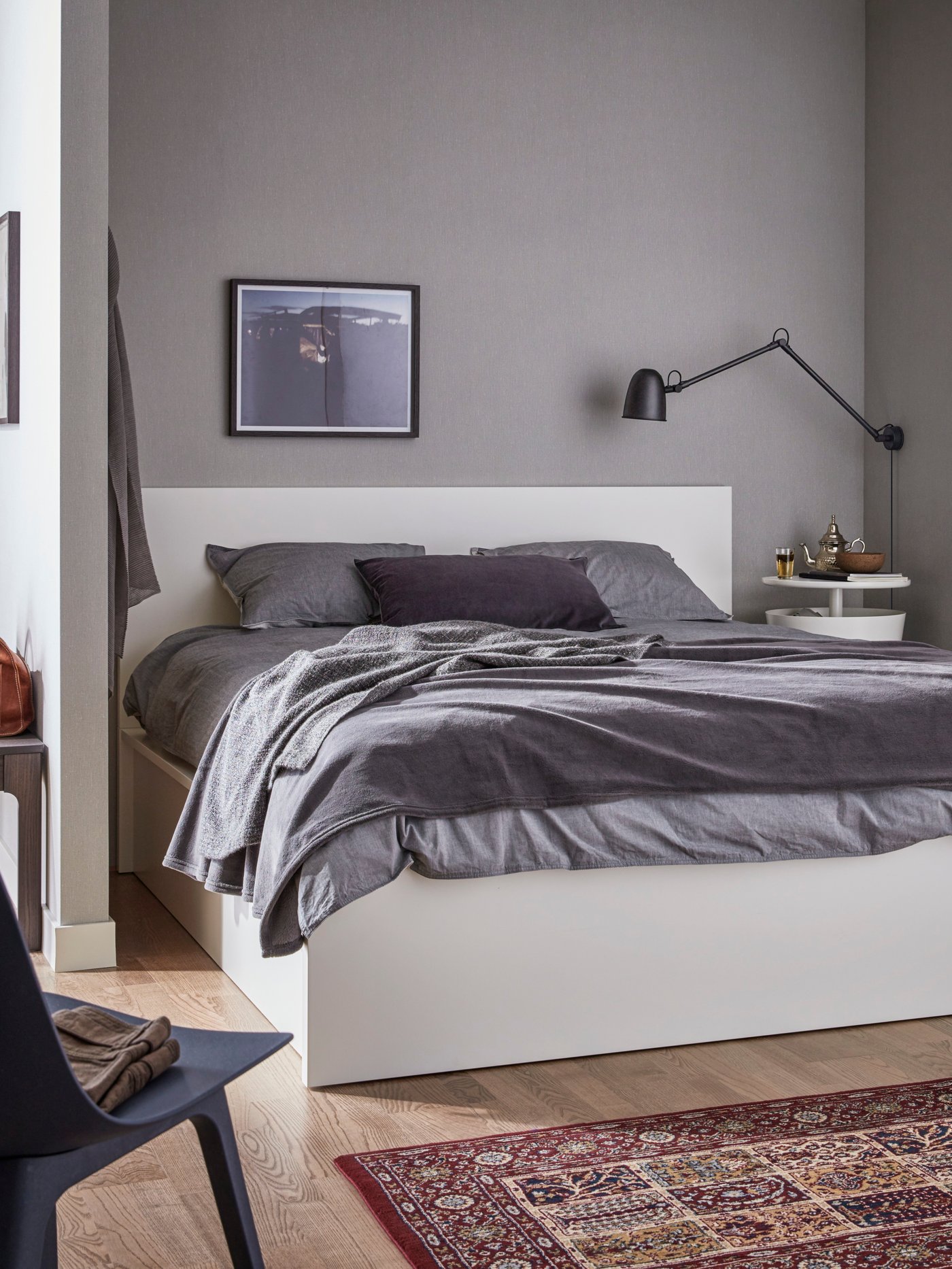 Cama otomana MALM de color banco con ropa de cama gris y una manta gris oscuro junto a una mesilla de noche blanca con una pared gris de fondo.