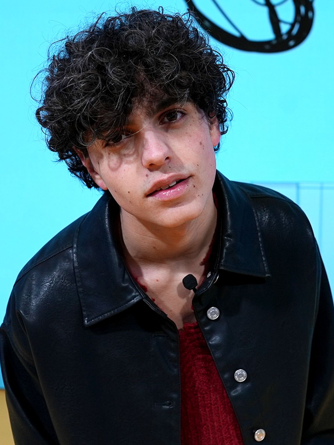 Niveo, ki nosi črno srajco in ogrlico, stoji na odru pred ozadjem modre barve z izdelki podjetja IKEA, za ozadje poskrbi digitalni zaslon, ki se med milanskim tednom oblikovanja 2024 uporablja na odru prizorišča IKEA. 