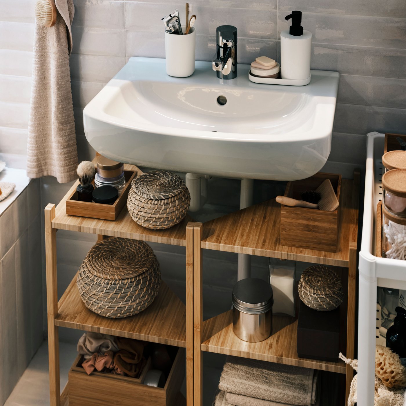 A small bathroom with two bamboo RÅGRUND corner shelves under a sink with baskets and boxes filled with essentials.