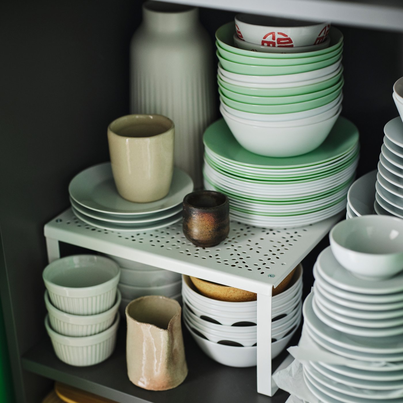 A white VARIERA shelf insert sits in a shelving unit. There are stacks of bowls and plates on, under, and next to it.