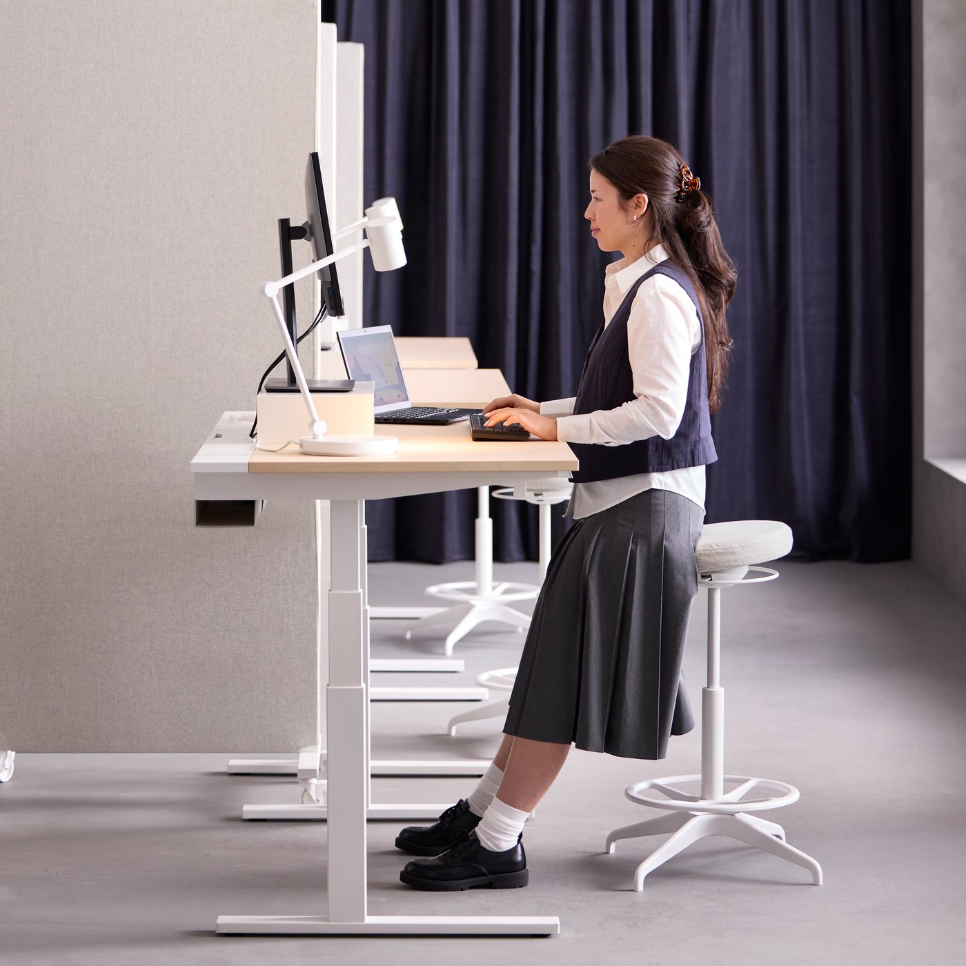 White MITTZON birch veneer sit/stand desk in mid-high mode with a person leaning on sit/stand support while working.