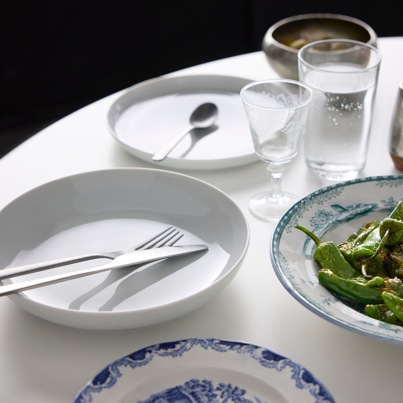 Part of a table set for a meal, with PRAKTBOTIA cutlery, an IKEA 365+ deep plate, an IKEA 365+ side plate, glasses and food.