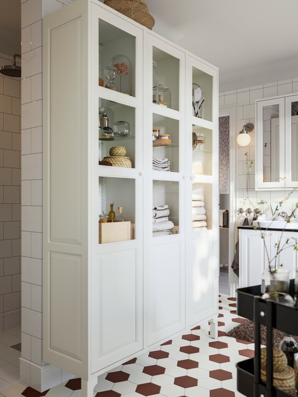 A bathroom featuring a white TÄNNFORSEN high cabinet with glass doors filled with towels, boxes and bottles.
