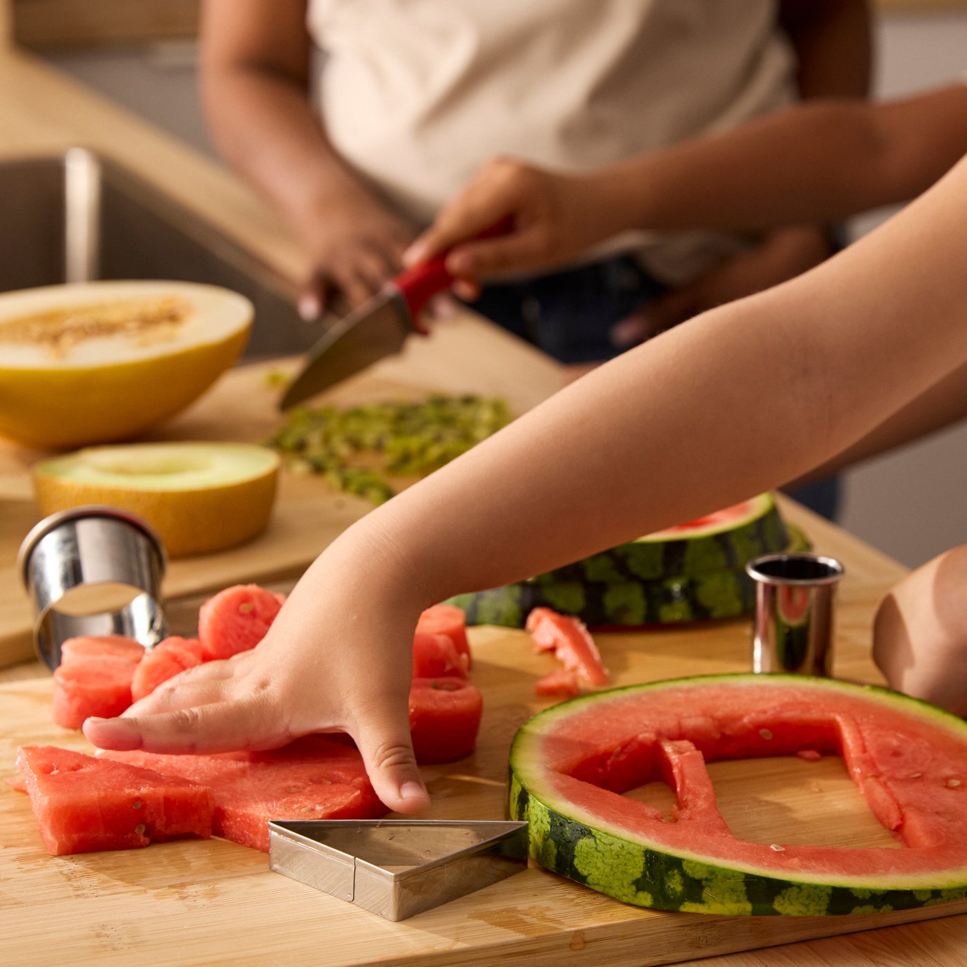 Close-up van kinderhandjes die meloen en watermeloen in stukjes snijden met LÄTTBAKAD uitsteekvormpjes.