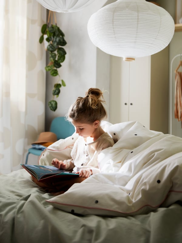 A girl reading on a bed with a dotted white/pink VÄNKRETS duvet cover and two white pendants above.