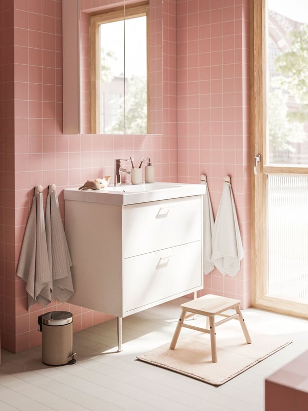 A white HAVBÄCK wash-stand with drawers and a mirror cabinet are placed in a bathroom with soft pink walls and a grey floor.