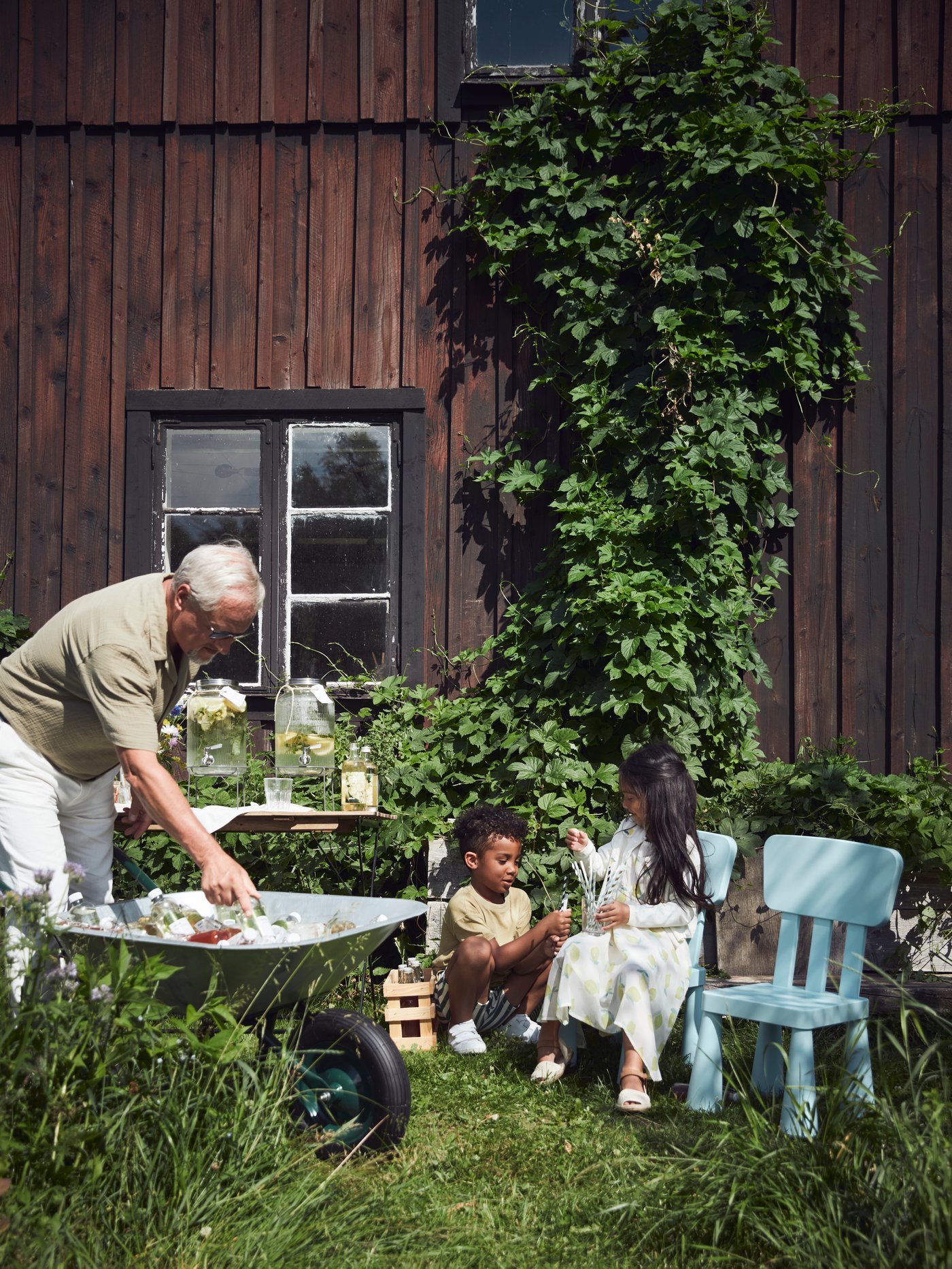 Dans un jardin, deux enfants discutent lors d’une fête d’été sur des chaises MAMMUT pendant que leur grand-père leur sert à boire.
