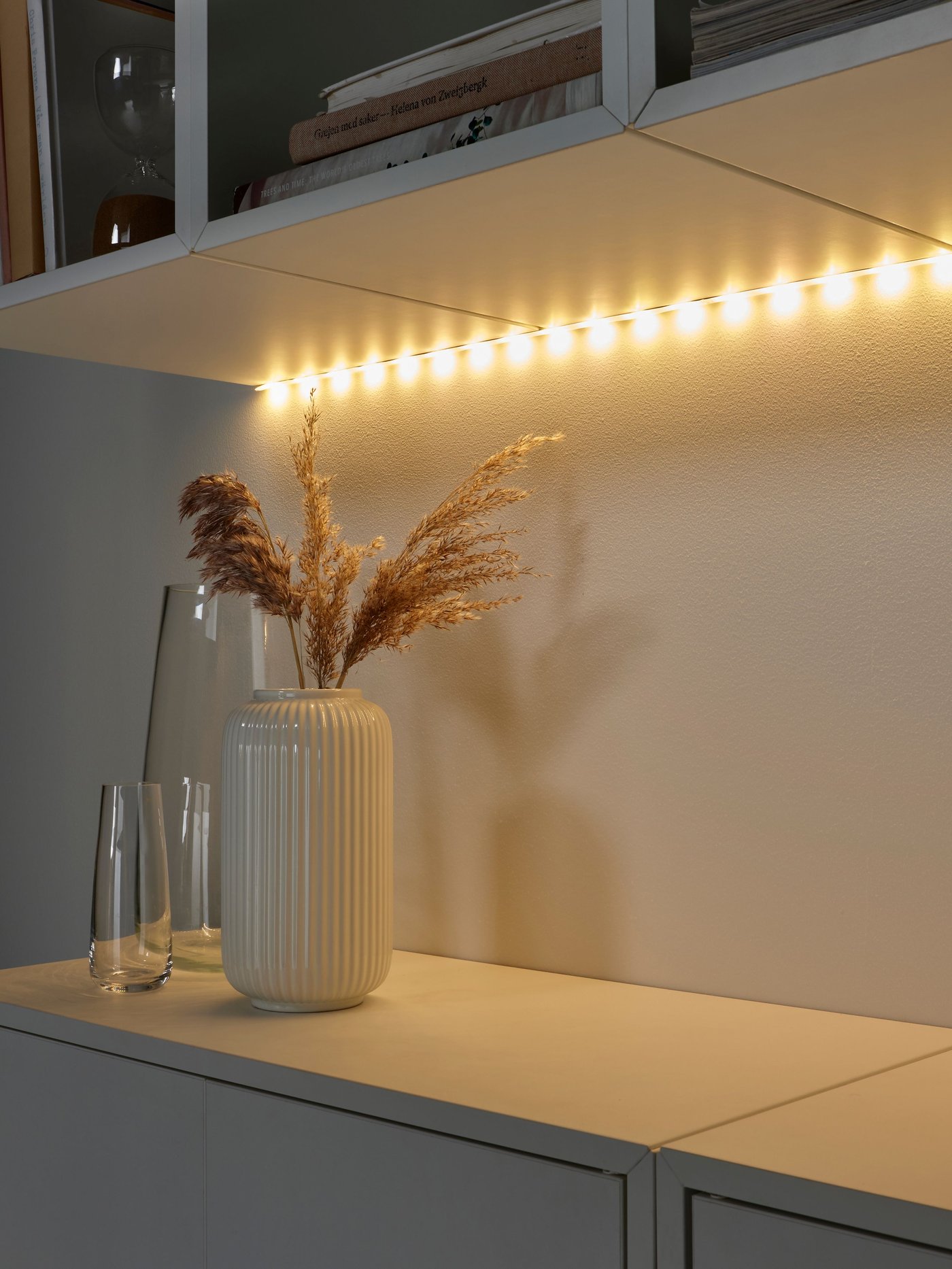 VATTENSTEN LED svetlobni trak je pritrjen na spodnjo stran stenskih omaric nad vazami, ki stojijo na drugih omaricah.