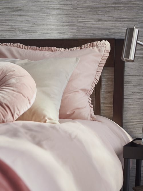 A KRANSBORRE cushion and another cushion with a SANELA cover lean against the headboard of a bed with pink KRANSKRAGE bed linen.