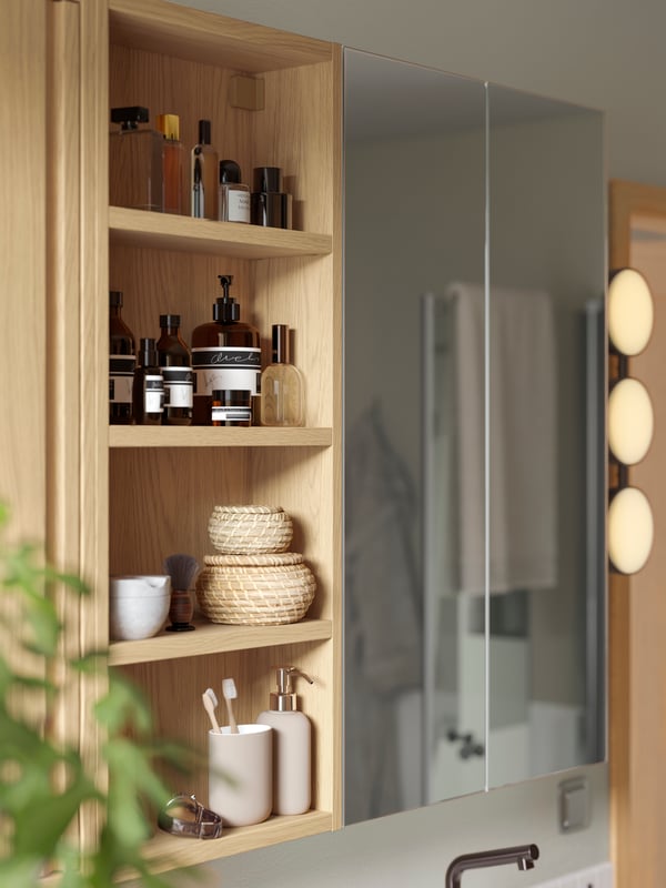 An open HAGAÅN wall cabinet with oak effect and a LETTAN mirror cabinet with door are next to a KABOMBA LED wall lamp.