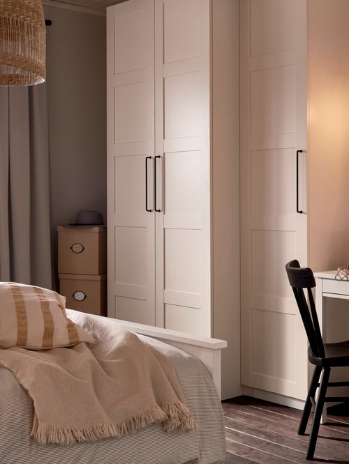 A white PAX wardrobe with BERGSBO doors stands near the foot of a white HEMNES bed and a black NORRARYD chair.