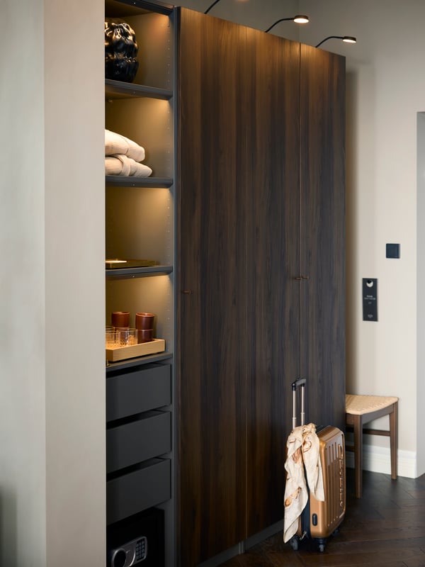 A PAX wardrobe with dark brown stained oak effect STORKLINTA doors is in a hotel room with a small suitcase in front of it.
