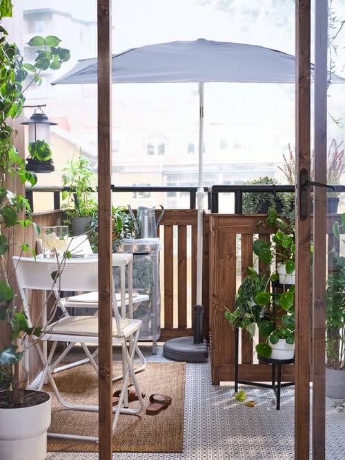 A small, leafy balcony with a white table and two chairs on top of a rug beside a grey LILLEÖ parasol in a BRAMSÖN stand.