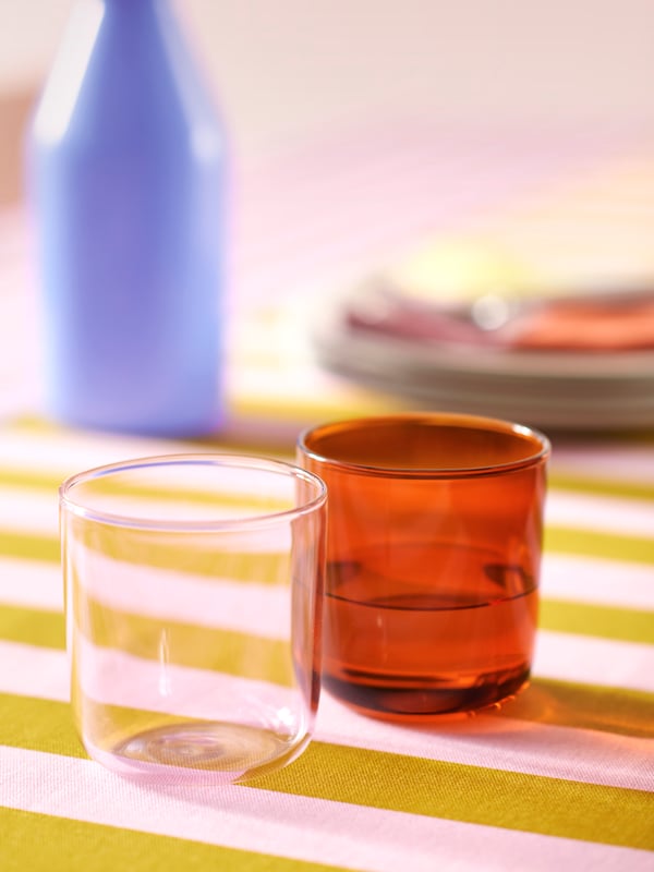 Two TESAMMANS glasses, one light pink and one brown, are on a table with TESAMMANS pre-cut fabric used as a tablecloth.