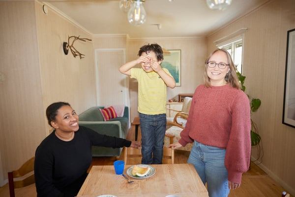 Alle, her wife and their son by their dining table, their son is standing on his chair covering his eyes and smiling.