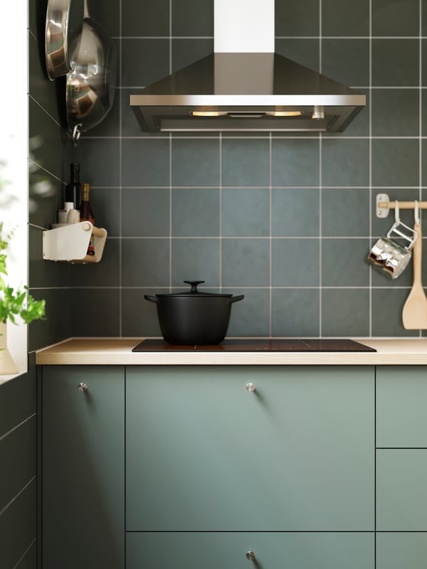 A green-tiled kitchen with NICKEBO fronts in matt grey-green with a hood, a black hob and a cast iron pot on top of it.