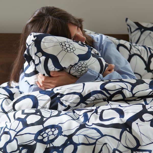 A person hugging a pillow while sitting in a bed with a white black/blue HÄRLIR duvet cover and matching pillowcases.