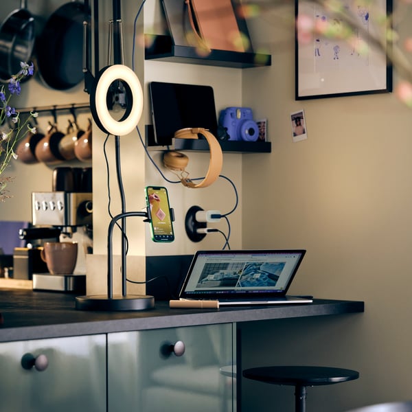 A shared charging space with a LÅNESPELARE ring light with phone holder, a laptop open and earphones hanging on the wall.