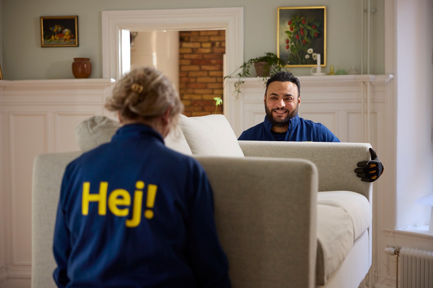 Two IKEA co-workers in blue uniforms carrying and positioning a sofa in a living room with a fireplace, artwork and plants in the background.