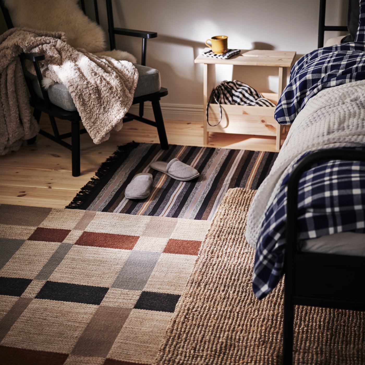 De vloer naast een bed is bedekt met drie verschillende tapijten in warme kleuren, met daarbovenop een paar TÅSJÖN pantoffels.