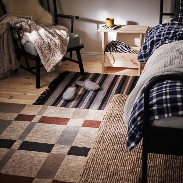 The floor next to a bed, covered with three different rugs in warm colors, topped off with a pair of TÅSJÖN slippers.