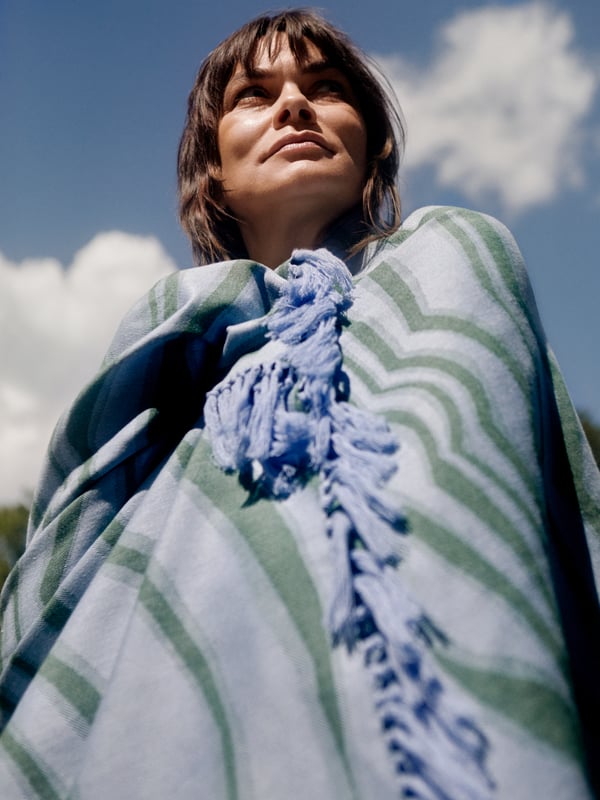 A woman smiles and stands close to an outdoor shower, with a blue and orange BASTUA bath sheet wrapped around her shoulders.
