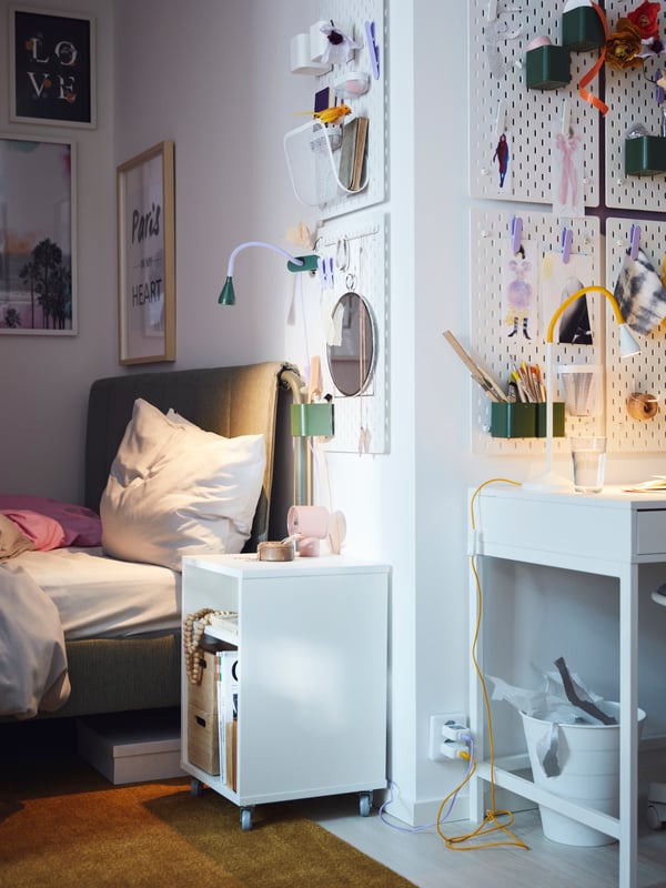 An inverted corner of a light room where a bed and VIHALS bedside table are on one side, and a white ALEX desk on the other.
