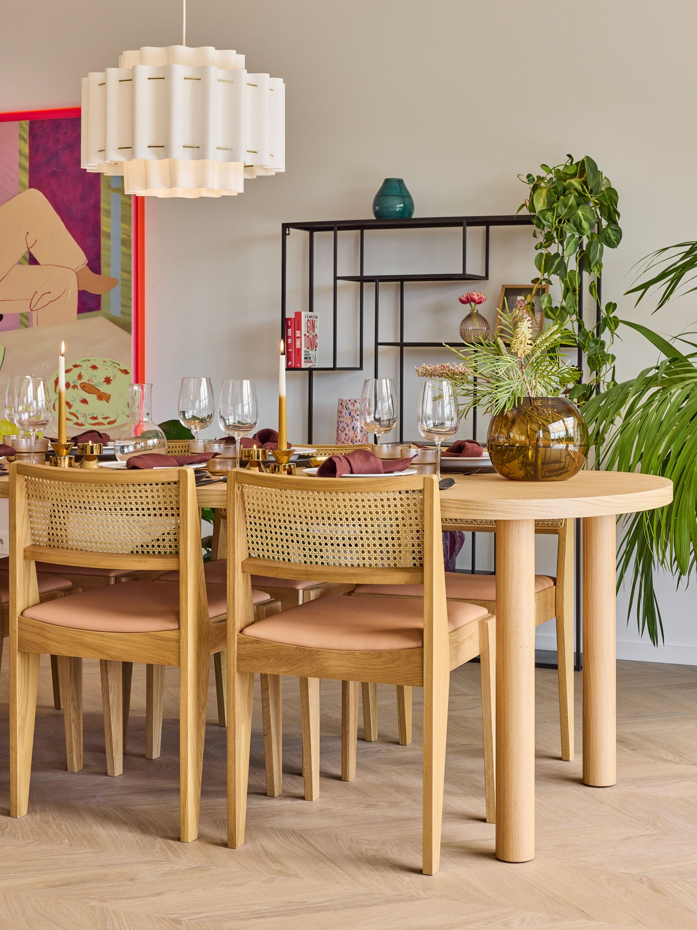 Gedekte eettafel met rotan stoelen, glazen en decoratie naast rek en kamerplanten.