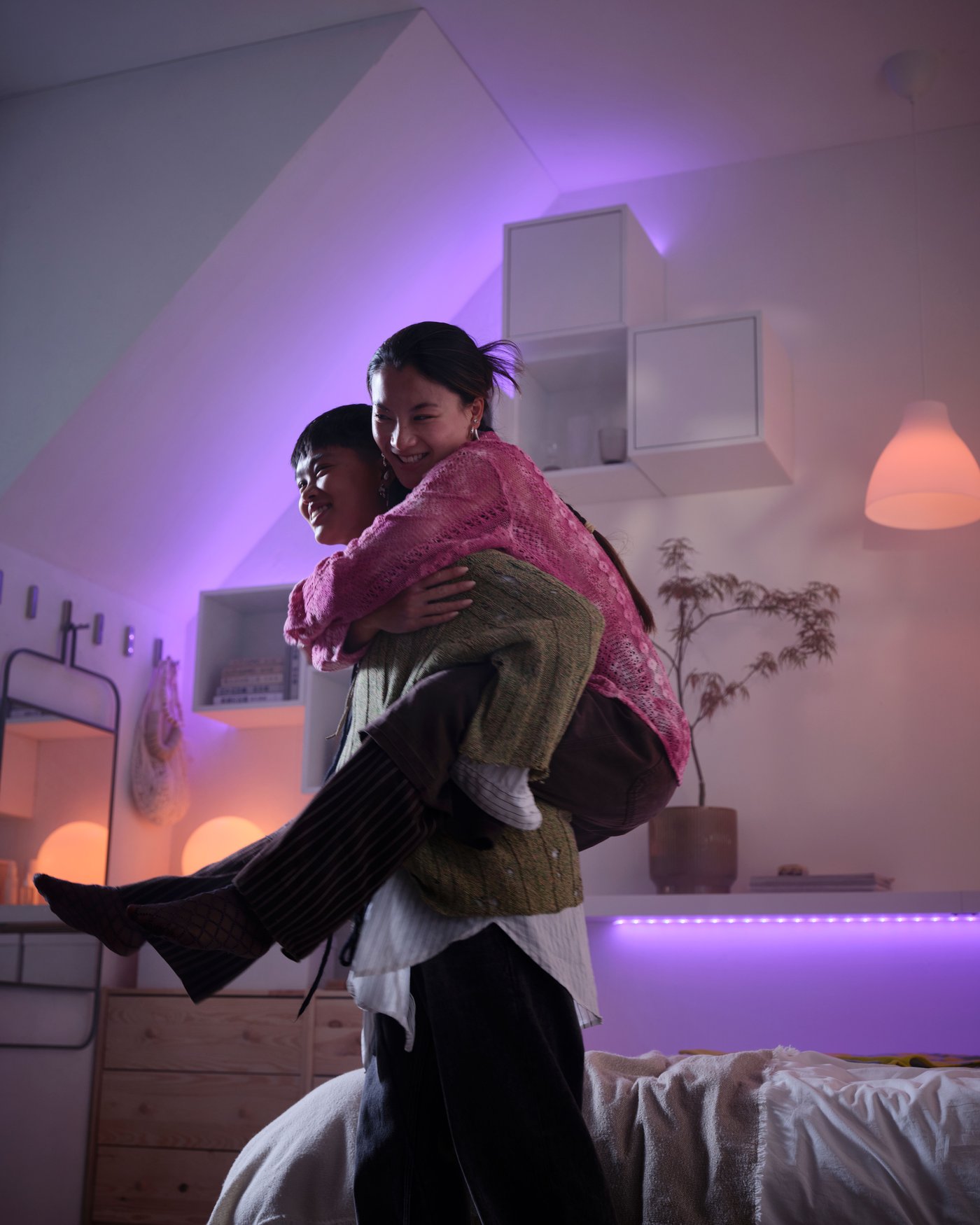 Due persone giocano in una stanza illuminata da strisce LED viola e rosa. Una persona porta l'altra sulle spalle, che sorride. Si vedono scaffali bianchi, un letto e uno specchio, con l'illuminazione a LED che crea un'atmosfera accogliente e divertente.