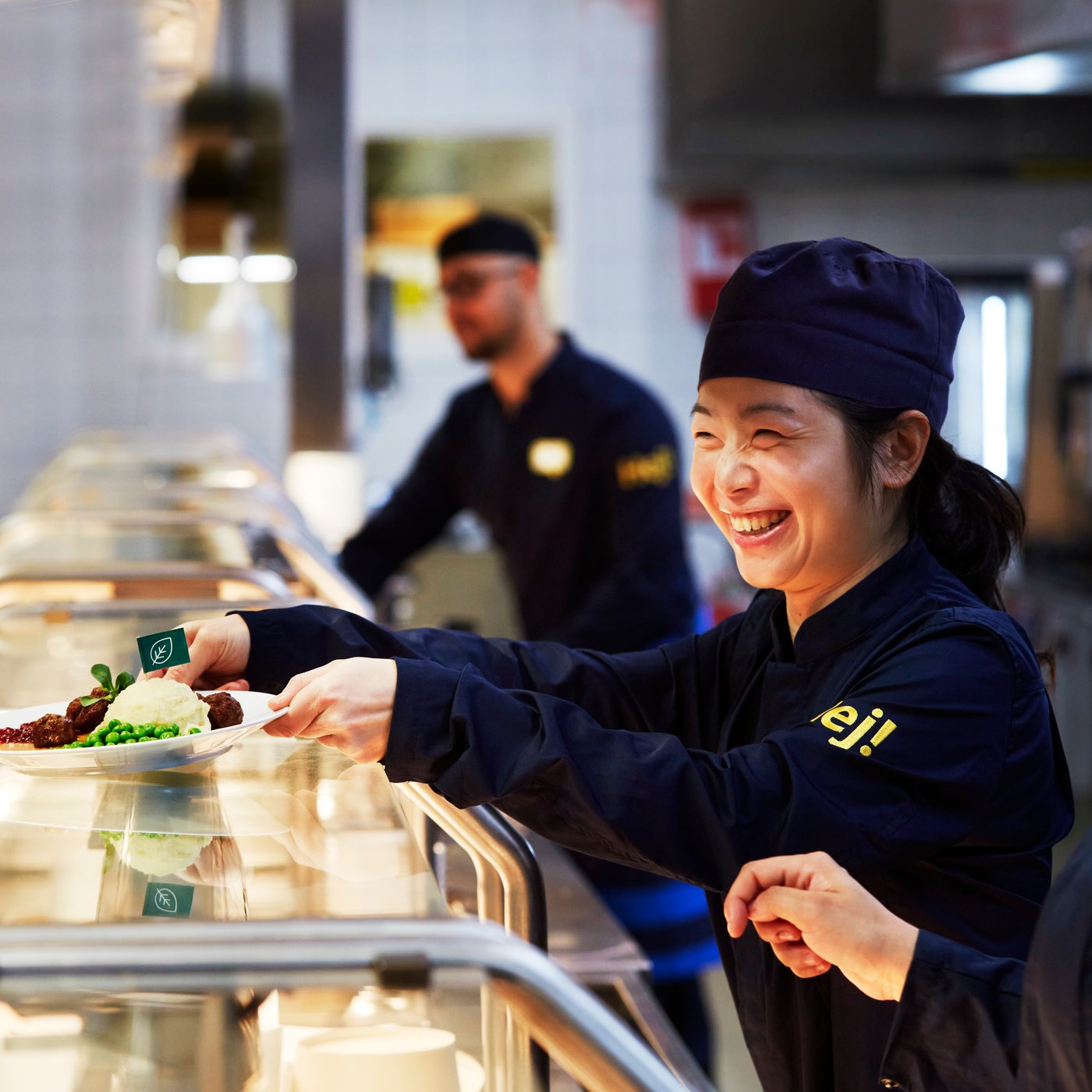 IKEA co-workers serving food at a cafeteria counter, wearing dark blue IKEA workwear, with kitchen staff and serving trays visible in the background.