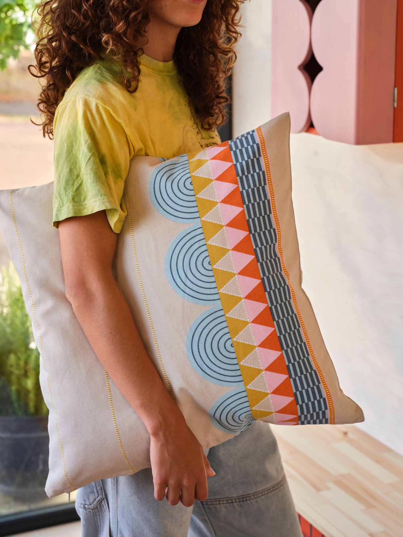 A person with a yellow shirt holds a cushion with a multicolour OMMJÄNGE cushion cover under their arm.