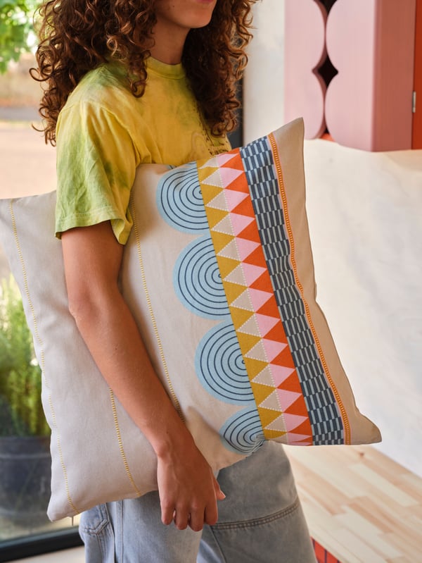 Una persona que porta una samarreta groga duu sota el braç un coixí amb una funda de coixí OMMJÄNGE de diversos colors.