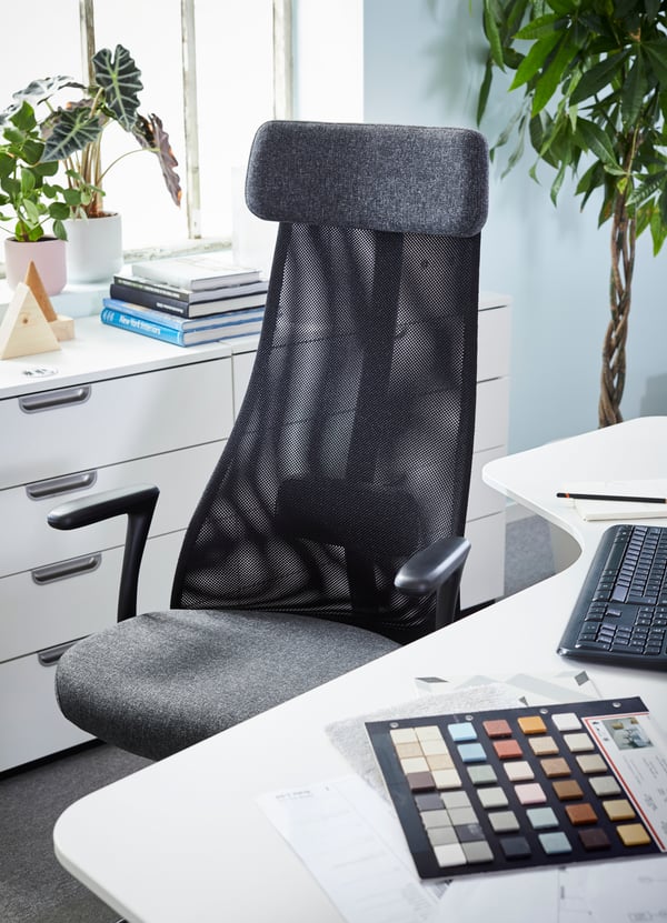 BEKANT white corner desk with a black mesh swivel office chair, by a window with potted plants and a pile of books.
