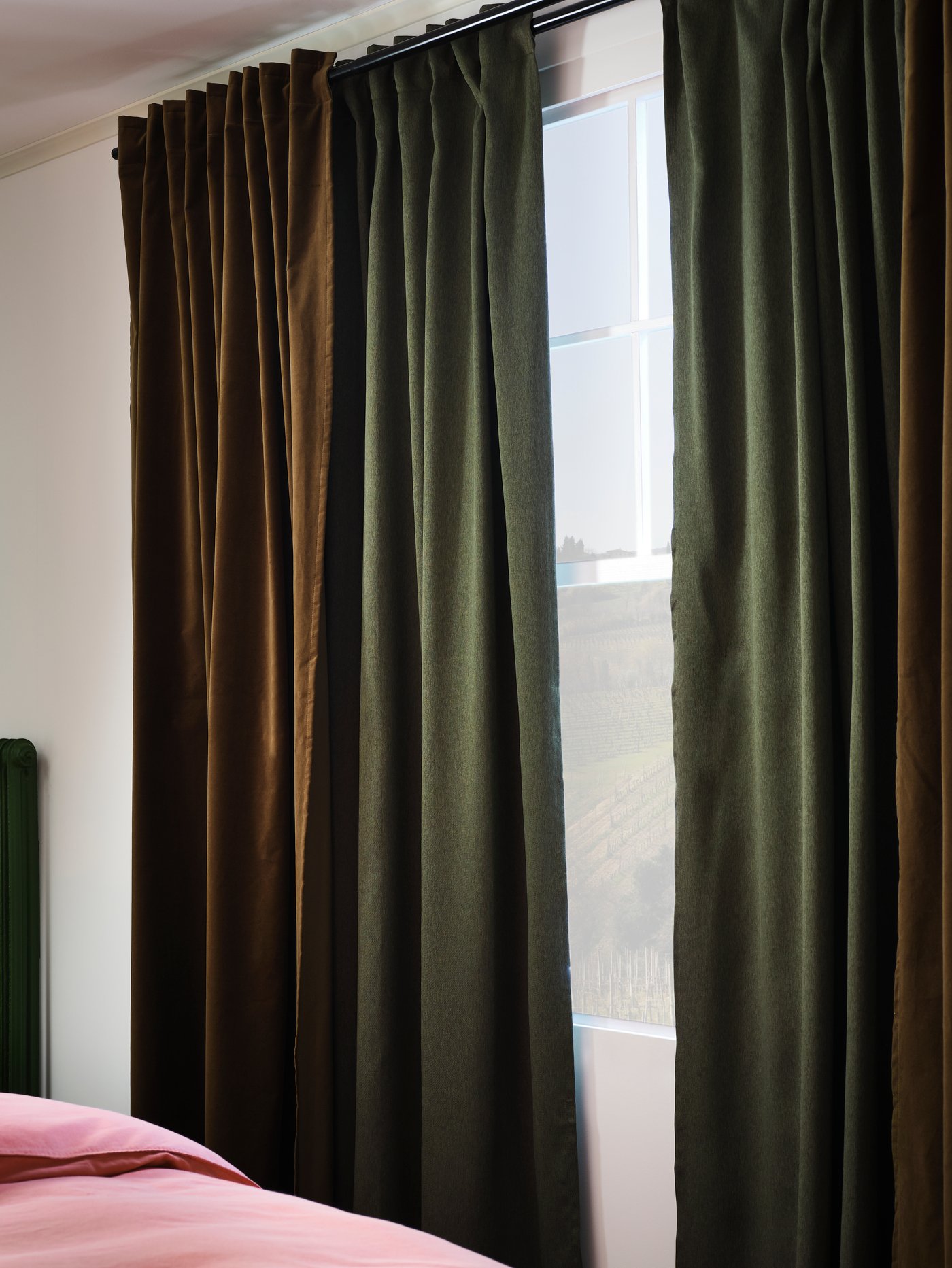 Okno se dvěma závěsy, na kterých visí zelené a olivově zelené závěsy. Vedle je postel se světle hnědočerveným povlečením.