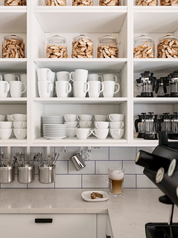 Part of a café interior with white, wall-mounted shelving filled with a neat arrangement of cups, mugs and jars.