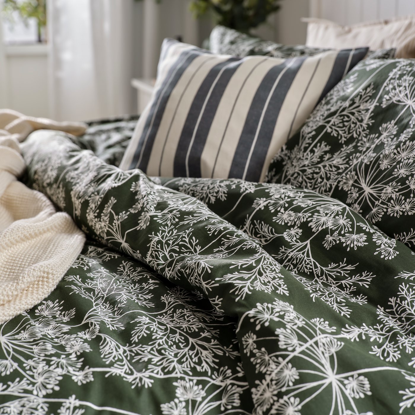 A close-up of a dark green/floral pattern DAGGBJÖRNBÄR duvet cover and pillowcases and KORALLBUSKE cushion cover on a bed.