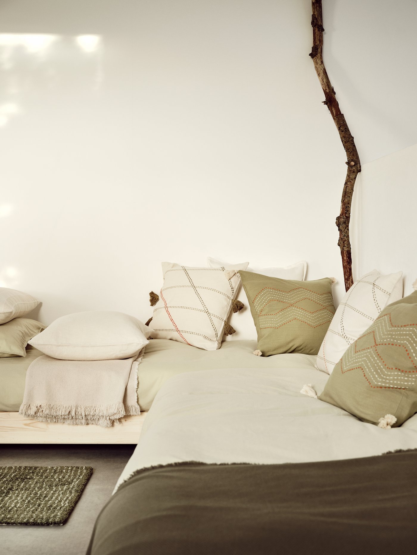 In the corner of a room, a cosy day bed is scattered with HERVOR and HALLVI cushion covers in off-white and green.
