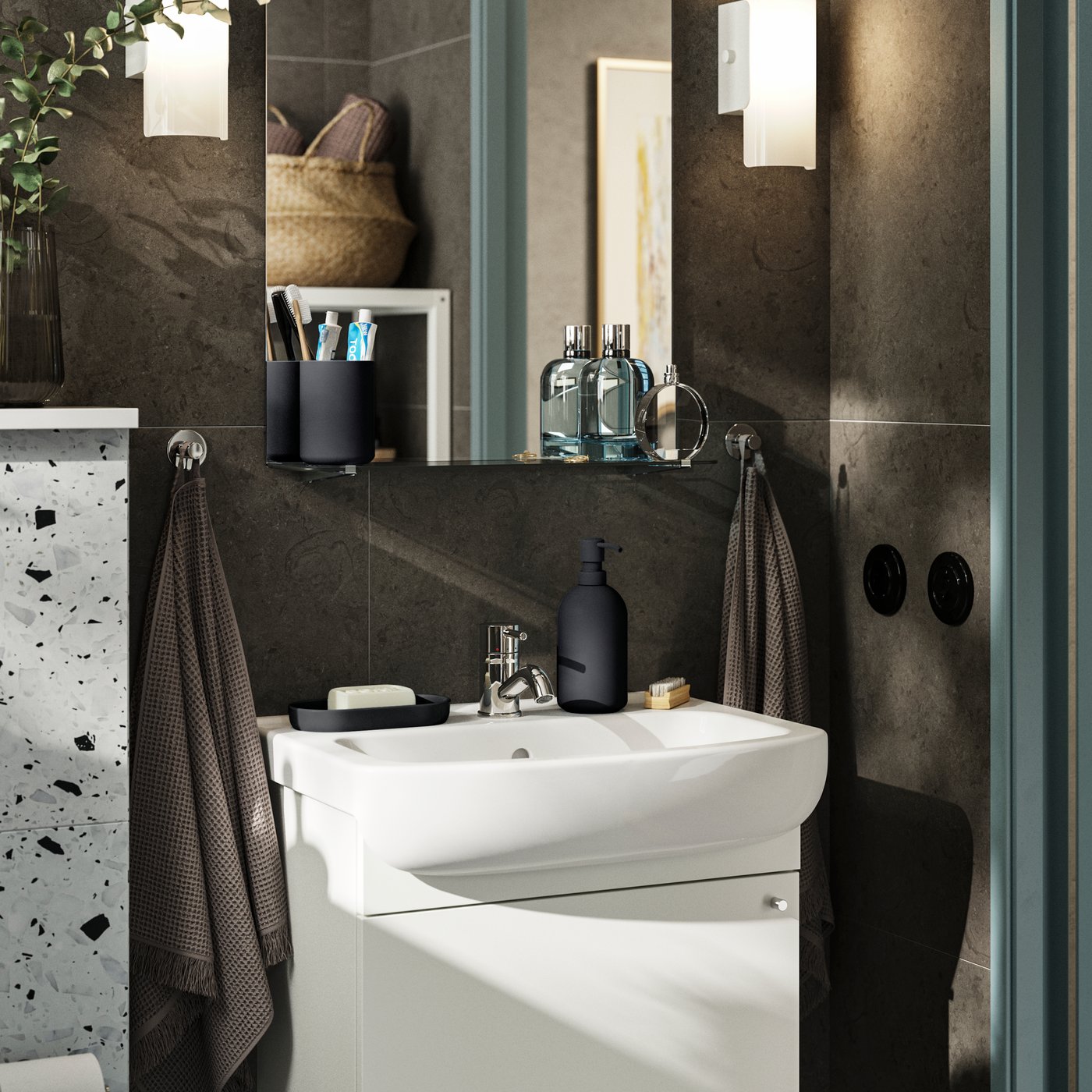 Majhna kopalnica s temno rjavimi stenami in belim pohištvom. NYSJÖN omarica z enojnimi vrati, za katerimi je prostor za shranjevanje pod umivalnikom.