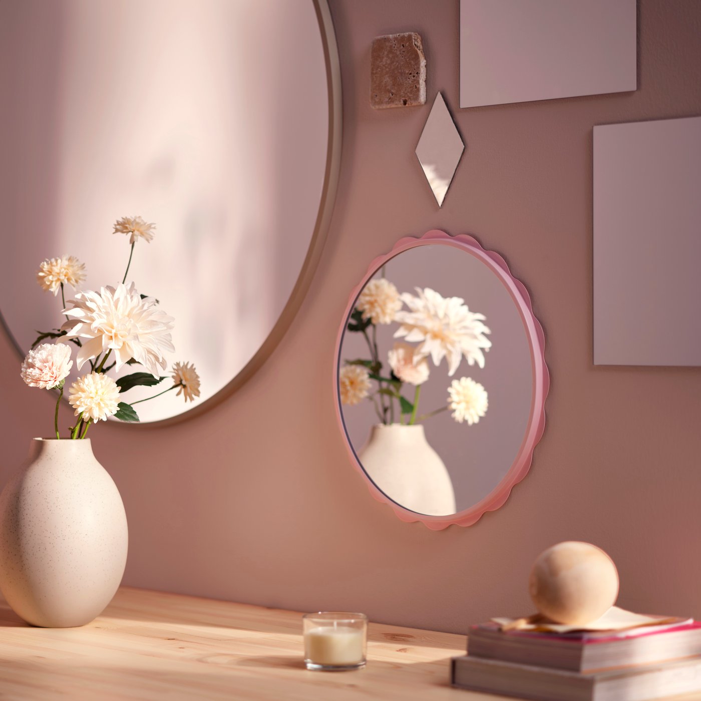 A round LÖNSÅS mirror reflecting a flower vase, hung above a wooden sideboard next to a larger KVALMSÖ mirror.