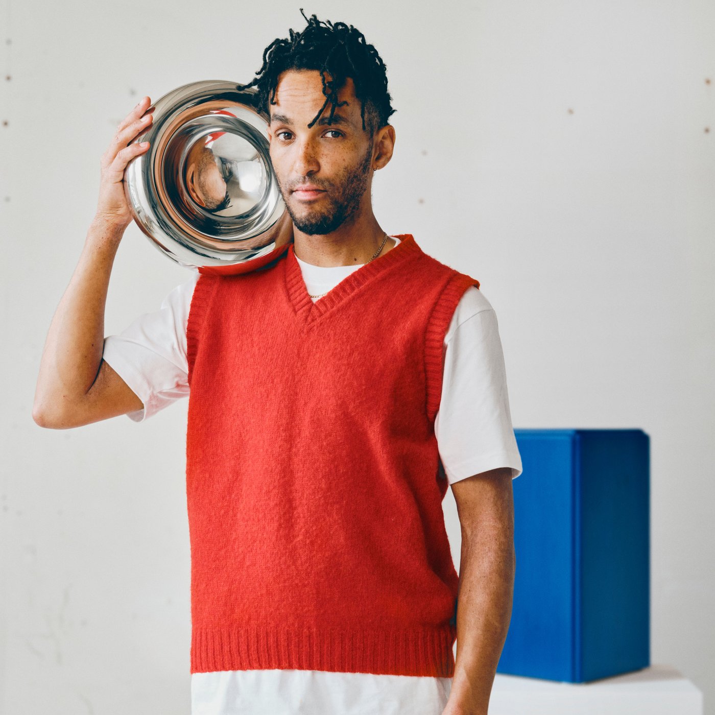 Andu Masebo holding a metallic bowl close to his head, behind him there’s one of his blue sculptures.