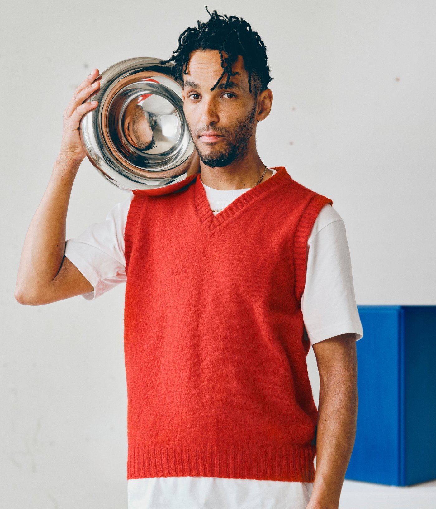 Andu Masebo holding a metallic bowl close to his head, behind him there’s one of his blue sculptures.