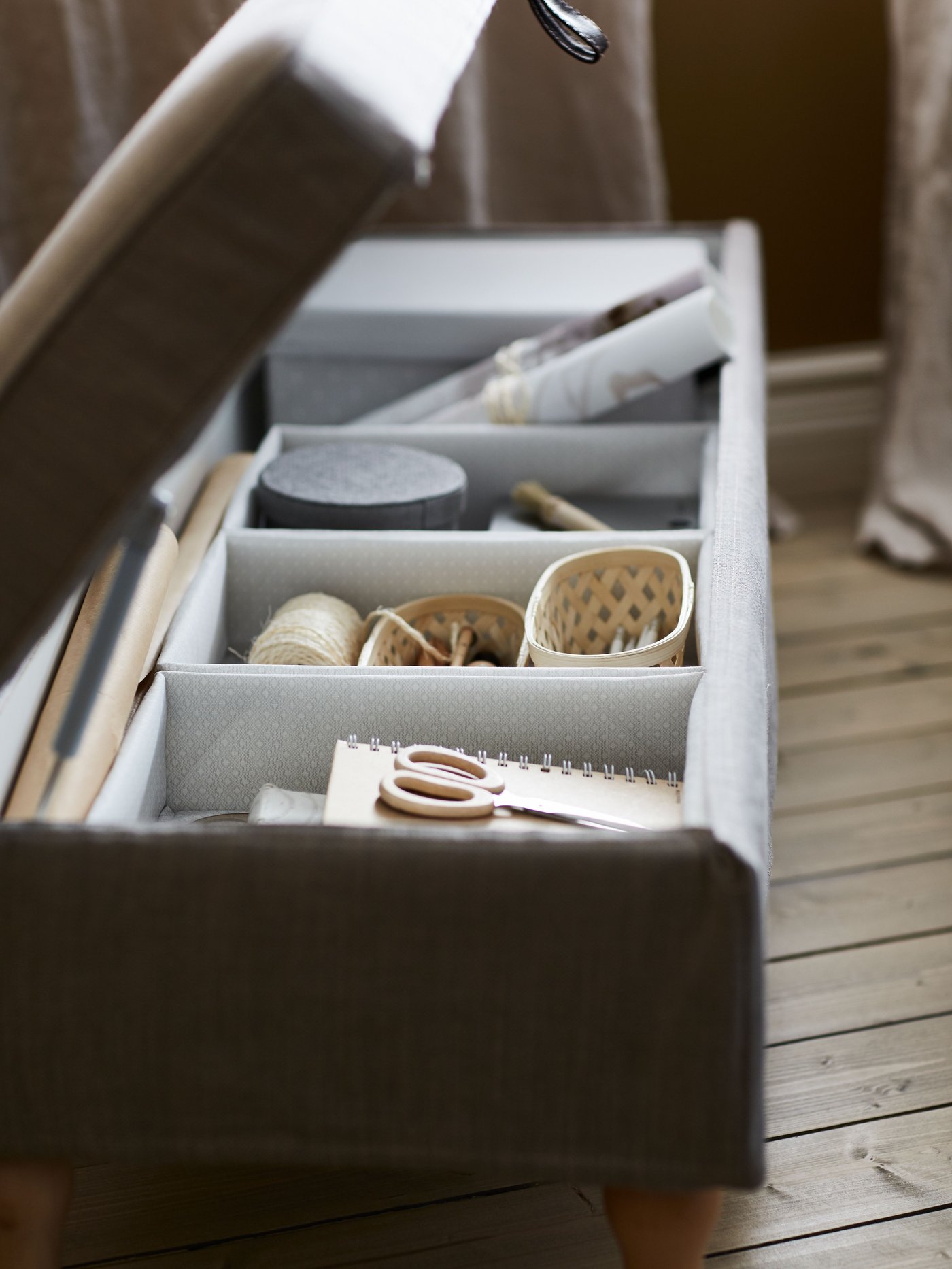 Suelo de madera con una caja de almacenaje gris con la tapa abierta. Hay varias cajas pequeñas, unas tijeras y material de costura dentro.