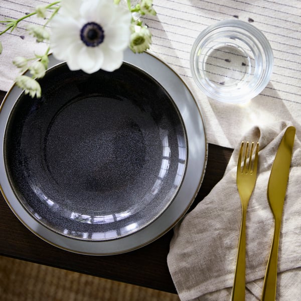 A dark grey GLADELIG deep plate sits on a patterned table with a glass of water, cutlery, and flowers nearby.