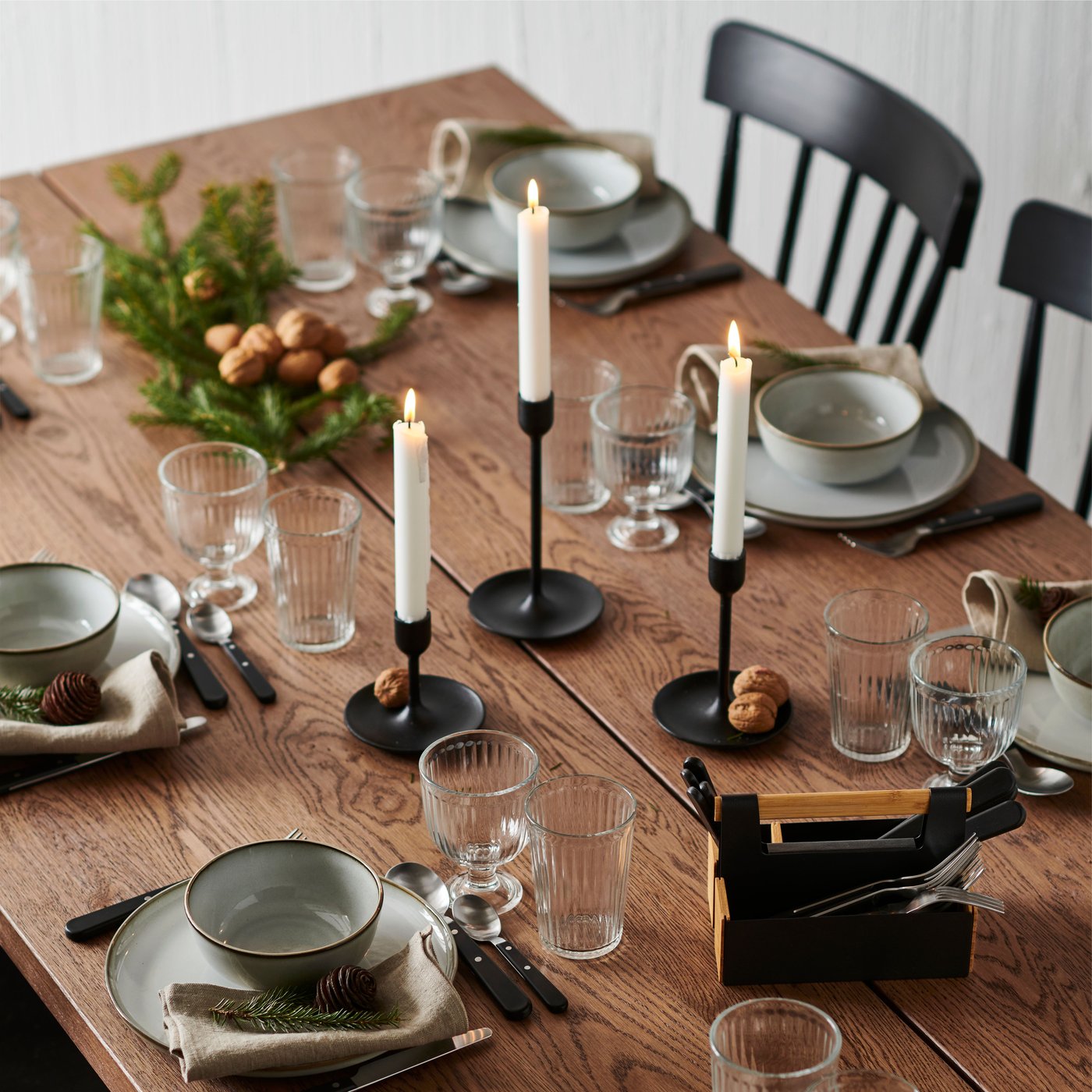 A wooden table set with GLADELIG crockery, glasses, napkins, candles, evergreen branches and nuts scattered.