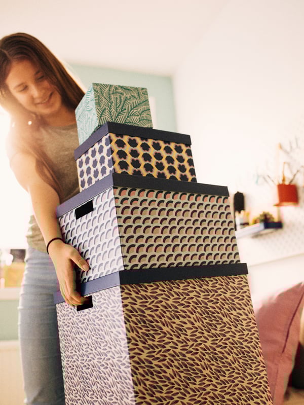 A pile of four TJENA storage boxes in different sizes and patterns, largest at the bottom, being picked up by a teen girl.