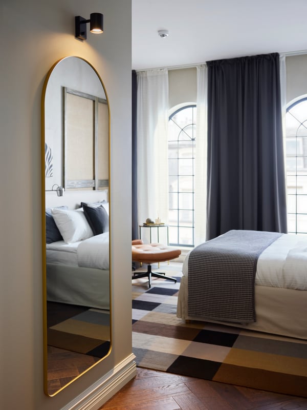 Under a lit wall lamp is a LINDBYN mirror with a gold-coloured frame that is rounded on top, partly reflecting a hotel bed.