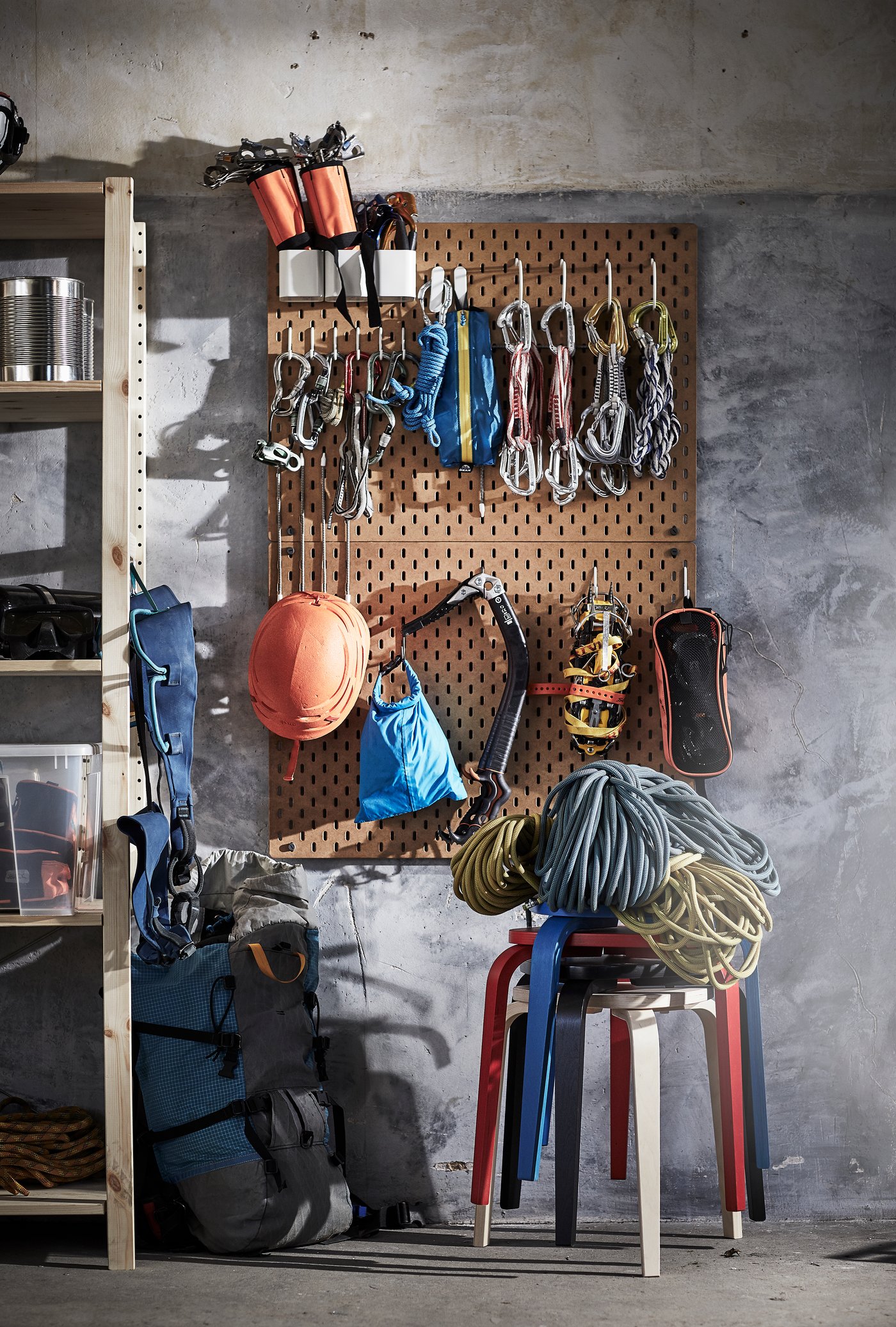 Stěna s perforovaným panelem IKEA SKÅDIS, na kterém visí horolezecké vybavení: lana, karabiny, popruhy, helma, rukavice a další doplňky. Pod panelem jsou složená horolezecká lana položená na barevných stoličkách IKEA FÄRGKLAR. Vlevo je dřevěný regál s úložnými boxy a vybavením.
