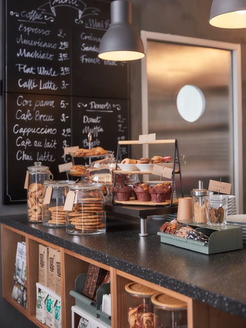 A serving tray, a box, jars, and a serving stand are on a black worktop, under which are beige wine shelves with items.
