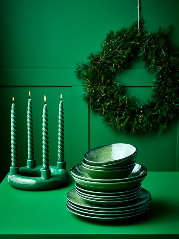 Gröna ljus i ljushållare bredvid gröna tallrikar på en grön hylla, med en julkrans hängande ovanför.
