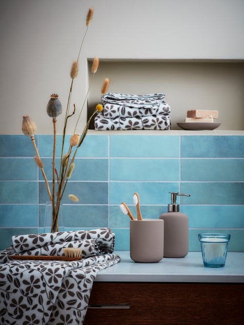 Zonă din baie cu faianță albastră, cu dozator de săpun și suport de periuță de dinți EKOLN, prosoape JUVELBLOMMA și obiecte decorative.